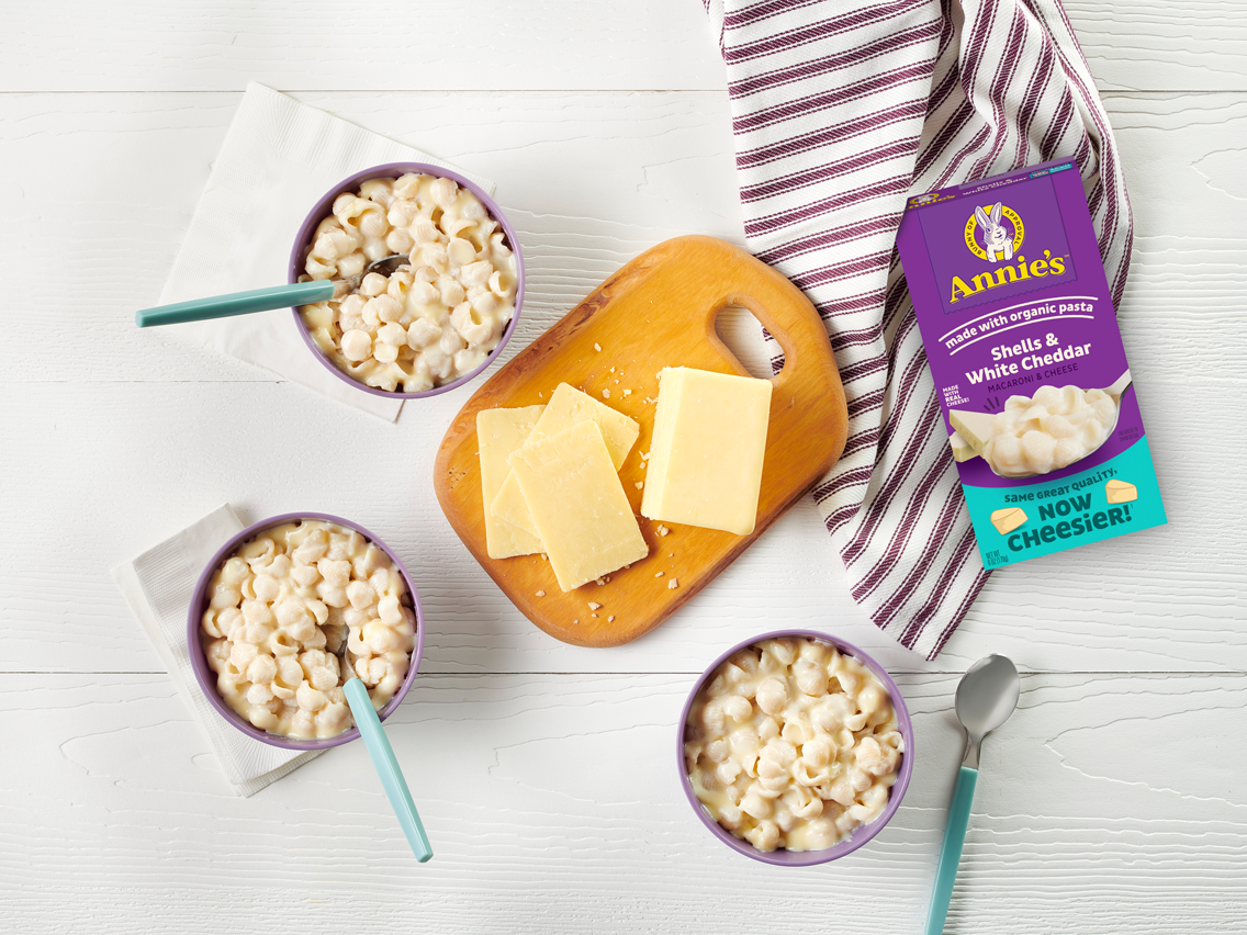 A box of Annie's shells & white cheddar macaroni & cheese, surrounded by three bowls of mac & cheese and a cutting board with slices of white cheddar