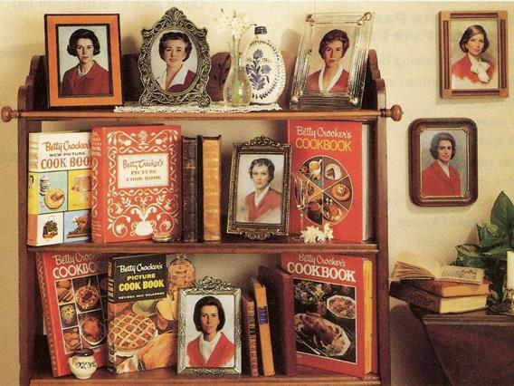 Three tier wood shelf in a kitchen displaying 6 Betty Crocker cookbook editions from 1950-1991, aside framed images of Betty Crocker headshots through the years.