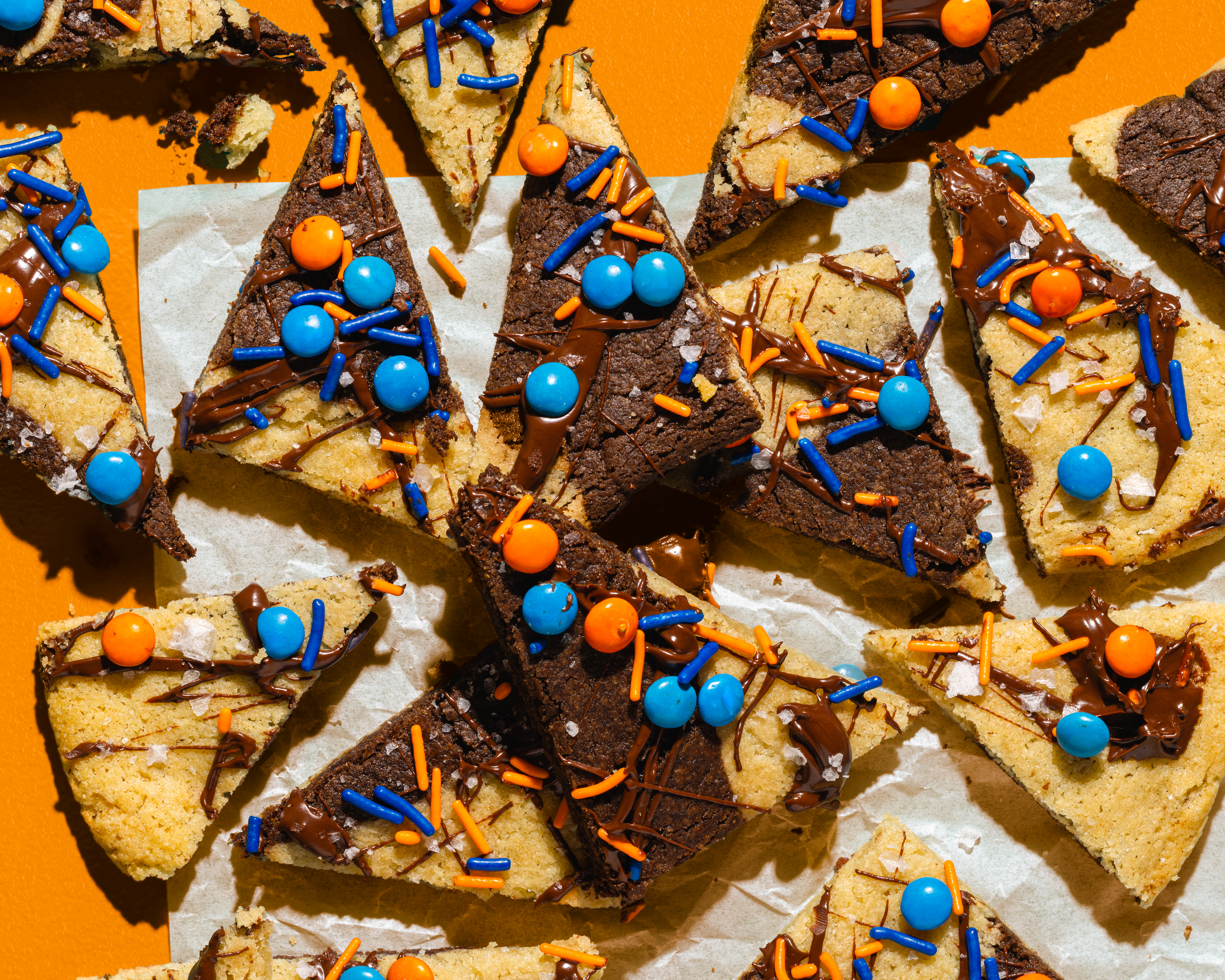 o	An overhead, close-up shot of triangular-shaped "Big Win Cookie Bark" dessert scattered on a piece of parchment paper against a solid orange background. The bars are made with marbled sugar cookie dough and cocoa powder, drizzled with melted chocolate and topped with blue and orange sprinkles, round candies, and a sprinkle of sea salt.
