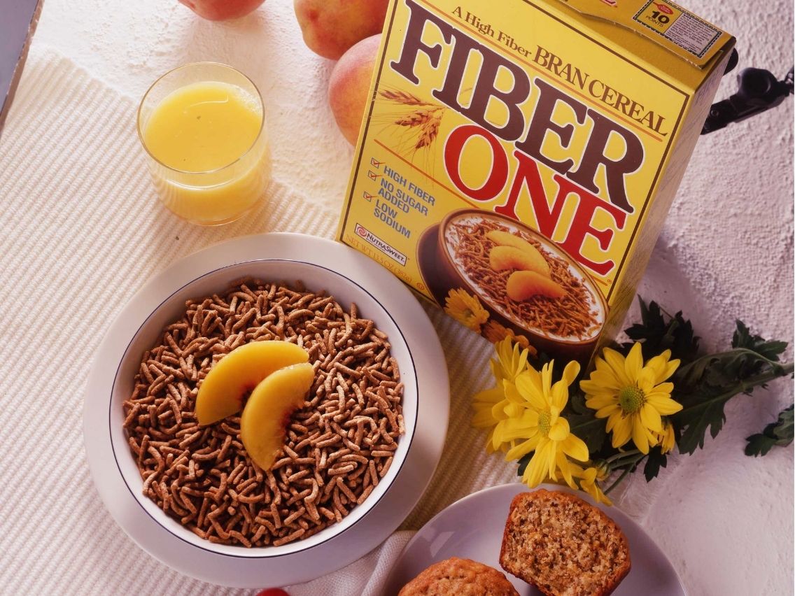 A bright, overhead shot of a breakfast setting on a white tablecloth. The scene includes a yellow box of Fiber One bran cereal, a white bowl filled with the cereal and garnished with two peach slices, a glass of orange juice, two bran muffins on a plate, and a small bouquet of yellow daisies.