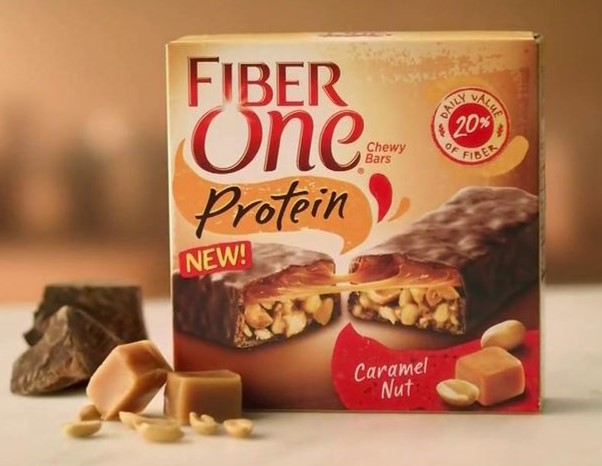 A close-up shot of a box of Fiber One Protein Chewy Bars in the 'Caramel Nut' flavor. The packaging shows a bar broken open to reveal a caramel and peanut filling. In the foreground, there are pieces of caramel, peanuts, and chocolate chunks.