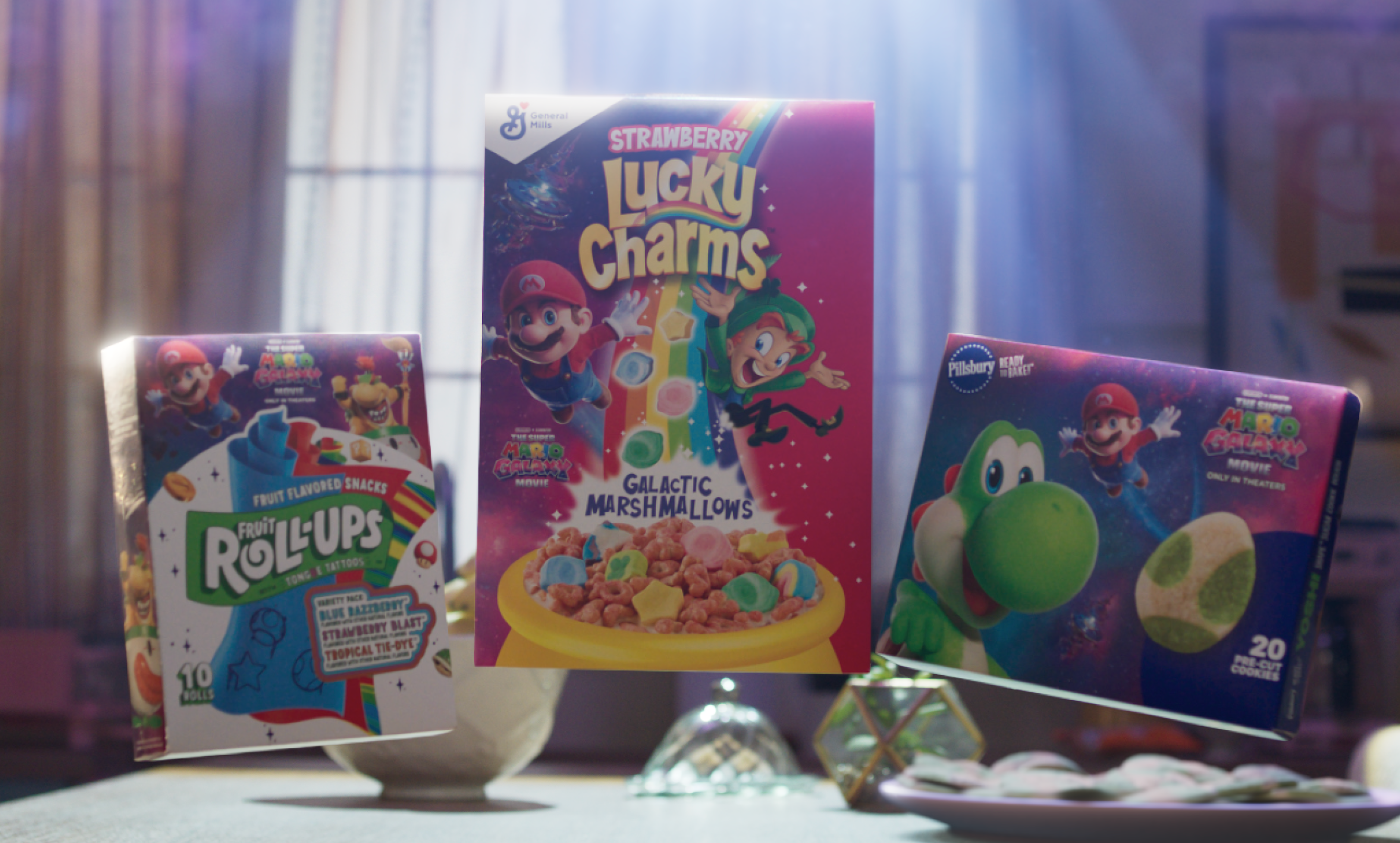 ​Lucky Charms Strawberry Cereal with Galactic Marshmallows, The Super Mario Galaxy Movie Edition, Pillsbury Ready to Bake! Yoshi Shape Sugar Cookie Dough and Fruit Roll-Ups Variety Pack, all in galactic-inspired packaging, float above a kitchen table with light shining in through a window in the background. 