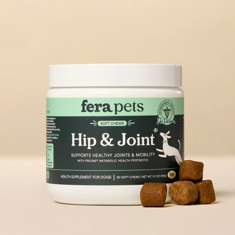 A white tub of Fera Pets Soft Chews Hip & Joint supplement for dogs, with a green label. Several brown, cube-shaped soft chews are scattered in front of the tub. The label features a cartoon dog jumping.