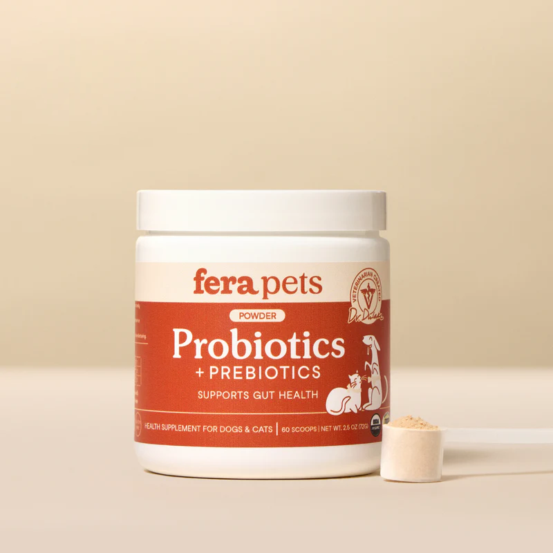 A white tub of Fera Pets Powder Probiotics + Prebiotics supplement, with a red label that states "Supports Gut Health." A small white scoop filled with the light-colored powder is placed in front of the tub. The label features cartoon illustrations of a dog and a cat.