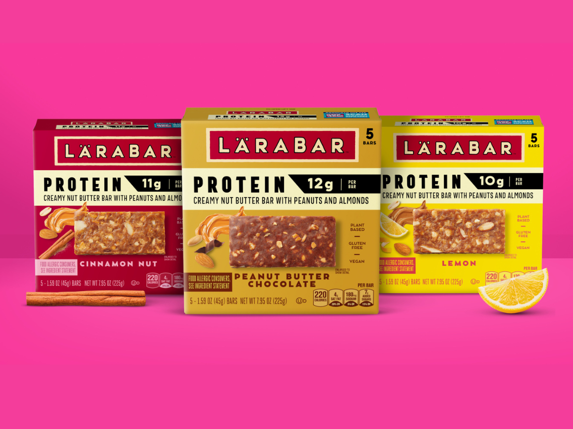  ​LÄRABAR Protein bars in Cinnamon Nut, Peanut Butter Chocolate and Lemon flavors displayed in colorful packaging against a bright pink background, highlighting the brand’s new plant-based protein bar lineup.