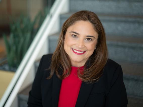Liz Mascolo, General Mills' Segment President, North America Pet, smiles warmly at the camera. She has brown hair and is wearing a black blazer over a red top, with a blurred background of stairs and greenery.