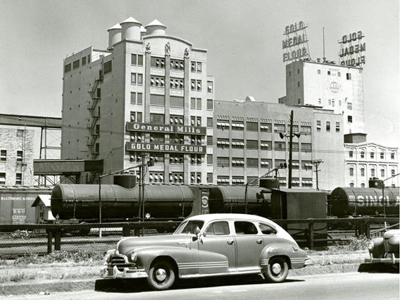 A black and white photograph shows the General Mills building with "General Mills" and "Gold Medal Flour" signs prominently displayed. On the roof of a taller building behind it, large signs spell out "GOLD MEDAL FLOUR." In the foreground, a vintage car is parked on the side of a road, and behind it, several train cars, including tank cars and a boxcar are visible on tracks.